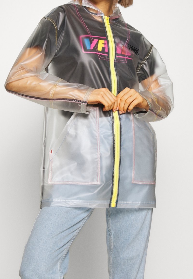 Imperméable en PVC transparent avec une fermeture éclair jaune, des accents roses et bleus, et un logo. Il présente des manches longues et deux poches avant.