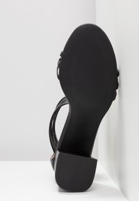 Sandale noire à talon haut avec semelle texturée et talon bloc, photographiée de dessous sur un fond d'angle blanc.