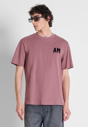 Giovane uomo con occhiali da sole e t-shirt malva con "AM" lettere sul petto, abbinata a jeans scuri su sfondo neutro.