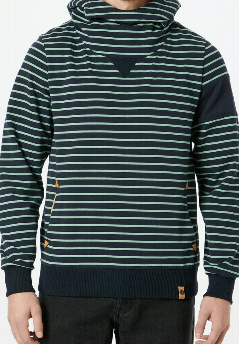 Homme portant un sweat à capuche à rayures horizontales bleu marine et vert avec un col montant et un pantalon foncé, faisant face à l'avant.