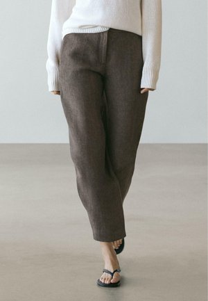 Personne portant un pantalon marron ample, un pull blanc et des sandales noires à lanières, marchant sur un sol clair devant un fond uni.