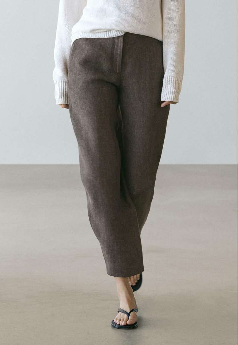 Personne portant un pantalon marron ample, un pull blanc et des sandales noires à lanières, marchant sur un sol clair devant un fond uni.