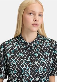 Zwarte blouse met een ruitpatroon in teal en wit, voorzien van een ruffles-kraag en korte mouwen, met een gladde textuur en een subtiele glans.