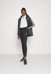 Chaqueta negra acolchada con rayas blancas, camiseta blanca con cremallera hasta el pecho y leggings negros con un logotipo. Zapatillas deportivas negras completan el conjunto.