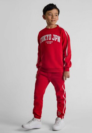 Red sweatshirt and joggers set featuring white "TOKYO JPN" text. Sweatshirt has long sleeves; pants have side stripes and logo at ankle.