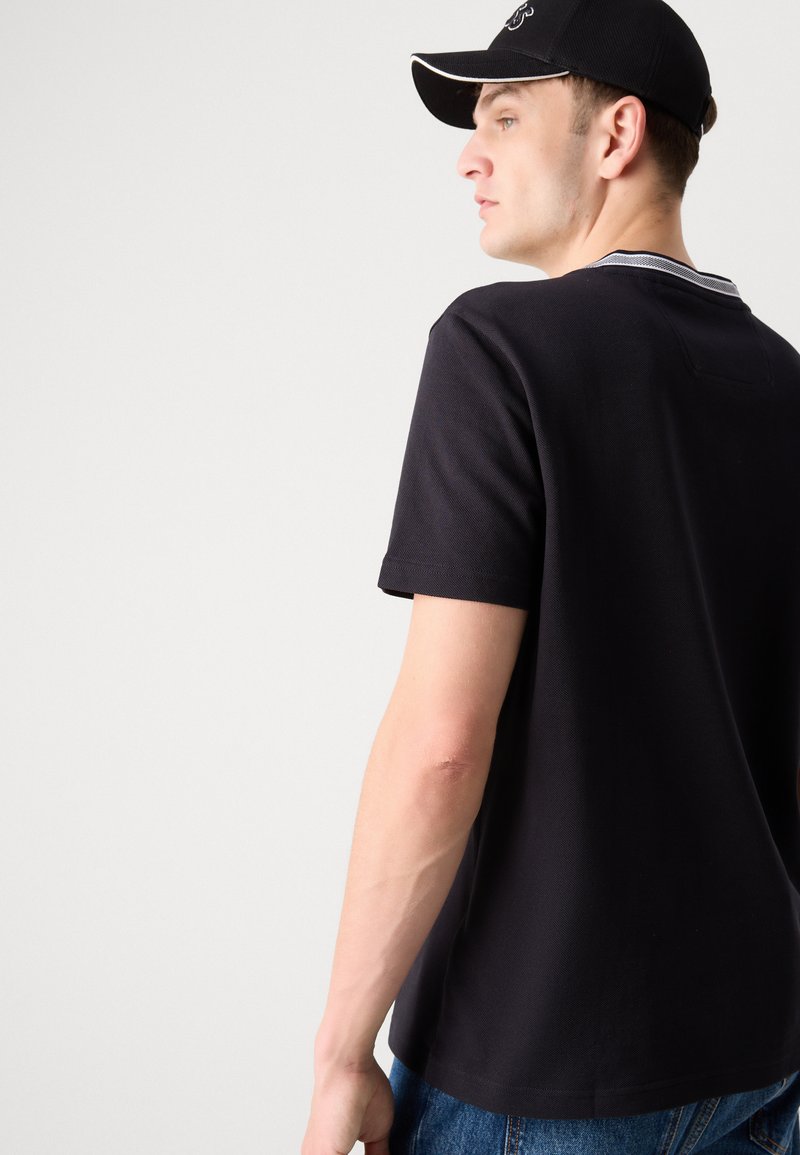 T-shirt noir à manches courtes en tissu texturé, col rond, avec de subtils détails à rayures blanches, associé à une casquette noire.