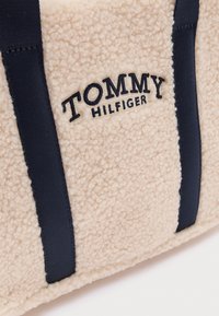 Beežikarva fliisest käekott mereväe siniste rihmadega, millel on tikitud "TOMMY HILFIGER" brändi nimi. Tekstuuriga pind annab pehme, hubase välimuse.