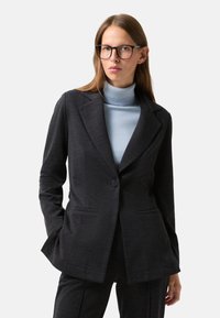 Veste taillée gris foncé avec un seul bouton, revers crantés, et deux poches avant, superposée à un pull col roulé bleu clair.