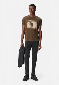 Camiseta de algodón marrón con un estampado gráfico, combinada con jeans oscuros y zapatos negros con cordones. Una chaqueta gris se sostiene en el brazo izquierdo.