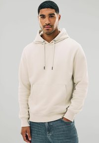 Sudadera de color beige claro hecha de una tela suave, con un bolsillo frontal, cordón ajustable, puños y dobladillo acanalados, combinada con jeans azules.