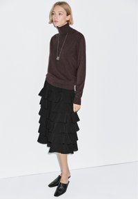 Brown turtleneck sweater paired with a black tiered skirt featuring ruffled layers. Black mules complete the outfit. Simple, modern design.