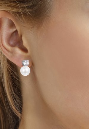 Primo piano di un orecchio che indossa un orecchino a bottone in argento con una pietra preziosa trasparente e una perla bianca pendente.