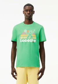 Camiseta de algodón verde con un gráfico de un jugador de tenis y el logo de "Lacoste" en blanco; cuello redondo y mangas cortas.