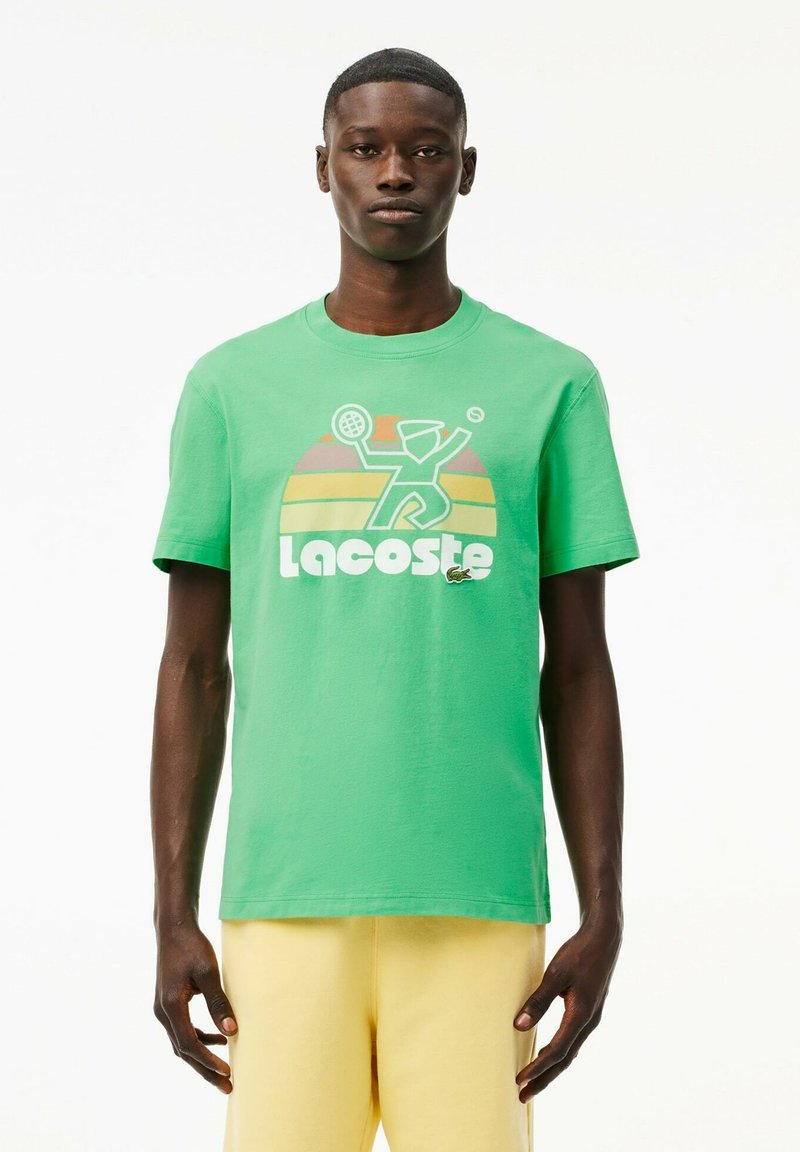 Camiseta de algodón verde con un gráfico de un jugador de tenis y el logo de "Lacoste" en blanco; cuello redondo y mangas cortas.