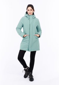 Mintgröna isolerade jacka med huva, dragkedjestängning och två framfickor, bärs över svarta byxor och vandringsskor. Slät textur.