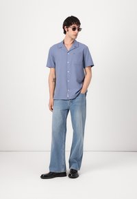 Lys blå skjorte med korte ermer og brystlomme, kombinert med vide lyse denimjeans og svarte sko, med solbriller som tilbehør.