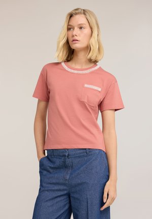 Kobieta nosząca różowy t-shirt z krótkim rękawem, z białym obszyciem na kołnierzu i kieszeni, w połączeniu z niebieskimi spodniami z wysokim stanem, stoi z jedną ręką w kieszeni.