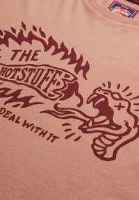 Camiseta rosa claro con un gráfico en rojo oscuro de una calavera en llamas que escupe fuego y texto "THE HOTSTUFF? DEAL WITH IT" en el pecho.