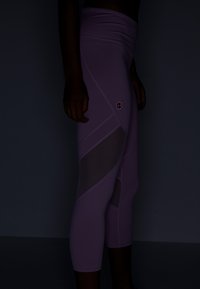 Ljusa lila leggings med hög midja, featuring meshpaneler och en liten logotyp i rosa. Designen inkluderar vinklade sömmar för en dynamisk look.