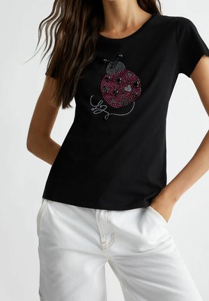 Vrouw draagt een zwart T-shirt met een rood-zwarte gestileerde lieveheersbeestje van juwelen, gecombineerd met een witte broek, staand met handen in de zakken.