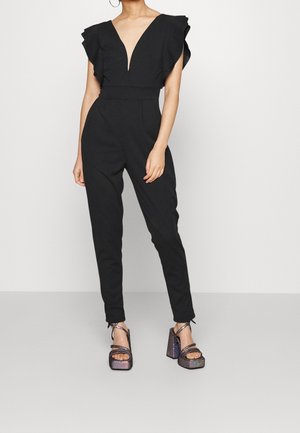 Vrouw draagt een zwarte mouwloze jumpsuit met gerimpelde schouders en een diepe V-hals, gecombineerd met sprankelende platform sandalen met hoge hak.