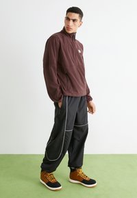 Bruine trui met een ritssluiting aan de voorkant en een zijzak, gecombineerd met zwarte, taps toelopende broek met witte stiksels, en bruine en zwarte enkellaarzen.