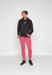 Svart hettegenser med lommer foran, parret med tettsittende rosa leggings. Personen har på seg svarte treningssko og holder et hoppetau.