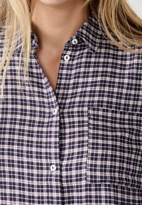 Koszula w niebiesko-białą kratę, zapinana na guziki, wykonana z lekkiego materiału, z kieszonką na piersi i klasycznym kołnierzem. Widoczne sześć guzików.