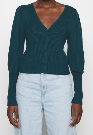 Cardigan en maille côtelée vert sarcelle foncé avec des boutons sur le devant, manches longues bouffantes, poignets ajustés, porté avec un jean taille haute bleu clair.