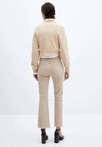Pull en beige clair, cropped, avec un col haut porté sur une chemise blanche. Jeans flare en beige clair, ajustés, avec des ourlets bruts et des chaussures à talons noires.