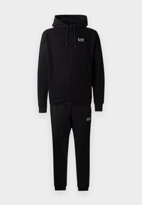 Črna zip-up kapuca s vrvicami in stranskimi žepi, v kombinaciji s črnimi trenirkami, ki imajo manšete in majhen logotip.