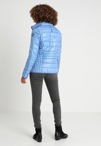 Hellblaues Puffer-Jacke mit horizontalem Steppmuster, hohem Kragen und Markenlogo, kombiniert mit grauen, taillierten Hosen und schwarzen Knöchelstiefeln.