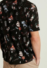 Korte mouwen shirt van zwarte stof met een kleurrijke print van tropische elementen, schedels, schepen en kaartomtrek met tekstaccenten.