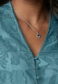 Blusa ricamata color teal con motivi floreali, scollatura a V e bottoni rotondi. Collana in argento con un pendente rettangolare.