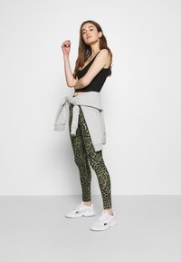 Sweatshirt cinza amarrado à cintura, top preto, leggings verde com padrão de leopardo, tênis brancos. Modelo exibe uma pose lateral.