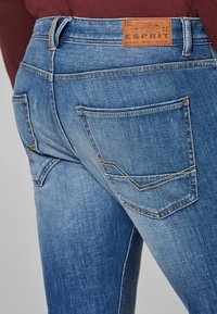 Lähikuva sinisistä farkuista, joissa on takataskut ja ruskea Esprit-brändin nahkalaatta vyötäröllä.