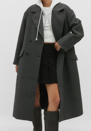 Femme portant un long manteau gris foncé par-dessus un sweat à capuche gris clair, une jupe noire et des bottes noires mi-mollet, debout devant un fond uni.