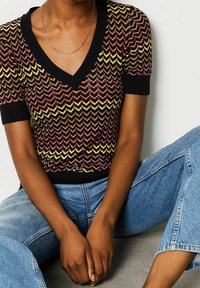 Neulepaita V-kaula-aukkolla, jossa on punaisen, keltaisen ja mustan siksak-kuvio. Lyhyet hihat ja vartalonmyötäinen malli. Yhdistetty vaaleansinisiin farkkuihin.