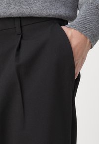 Osoba w czarnych spodniach i szarym swetrze wełnianym ma jedną rękę swobodnie włożoną w kieszeń spodni.