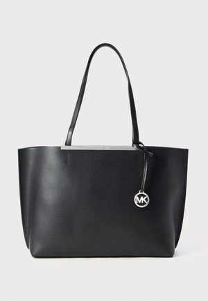 Schwarze Ledertasche mit doppelten Schulterriemen und einem silbernen, runden MK-Logoanhänger, der an einem Riemen hängt.