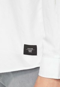 Biała koszula z długim rękawem z czarną metką materiałową z napisem "S.OLIVER ELEVATED CASUAL SINCE 1969" przeszytą w lewym dolnym rogu.