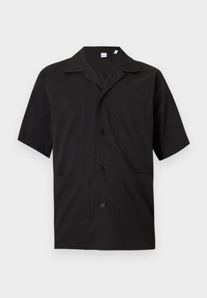 Man with short blonde hair wearing black short-sleeve buttoned shirt, olive green pants, and black shoes, standing against a plain white background.