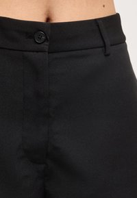 Pantalon noir taille haute avec fermeture à boutons et passants pour ceinture, montrant partiellement le torse et la taille sur un fond neutre.