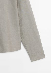 Manche longue d'une chemise en tissu tissé gris clair avec coutures visibles au niveau du poignet et de la couture latérale sur fond blanc.