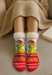 Gambe coperte con una coperta beige, indossando calze a tema Grinch rosse e bianche con un cappello di Babbo Natale e la scritta "Feelin' Grinchy", mentre si riposano su un divano.
