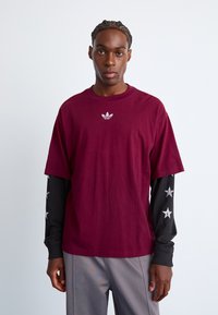 Bordeaux katoenen t-shirt met ronde hals en korte mouwen, voorzien van een klein wit logo op de borst. Zwarte lange mouwen shirt eronder met zilveren ster accents.