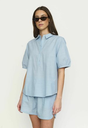 Nő világoskék rövid ujjú, galléros ingben és hozzá illő rövidnadrágban, sötét napszemüveget viselve, egyszínű háttér előtt pózol.