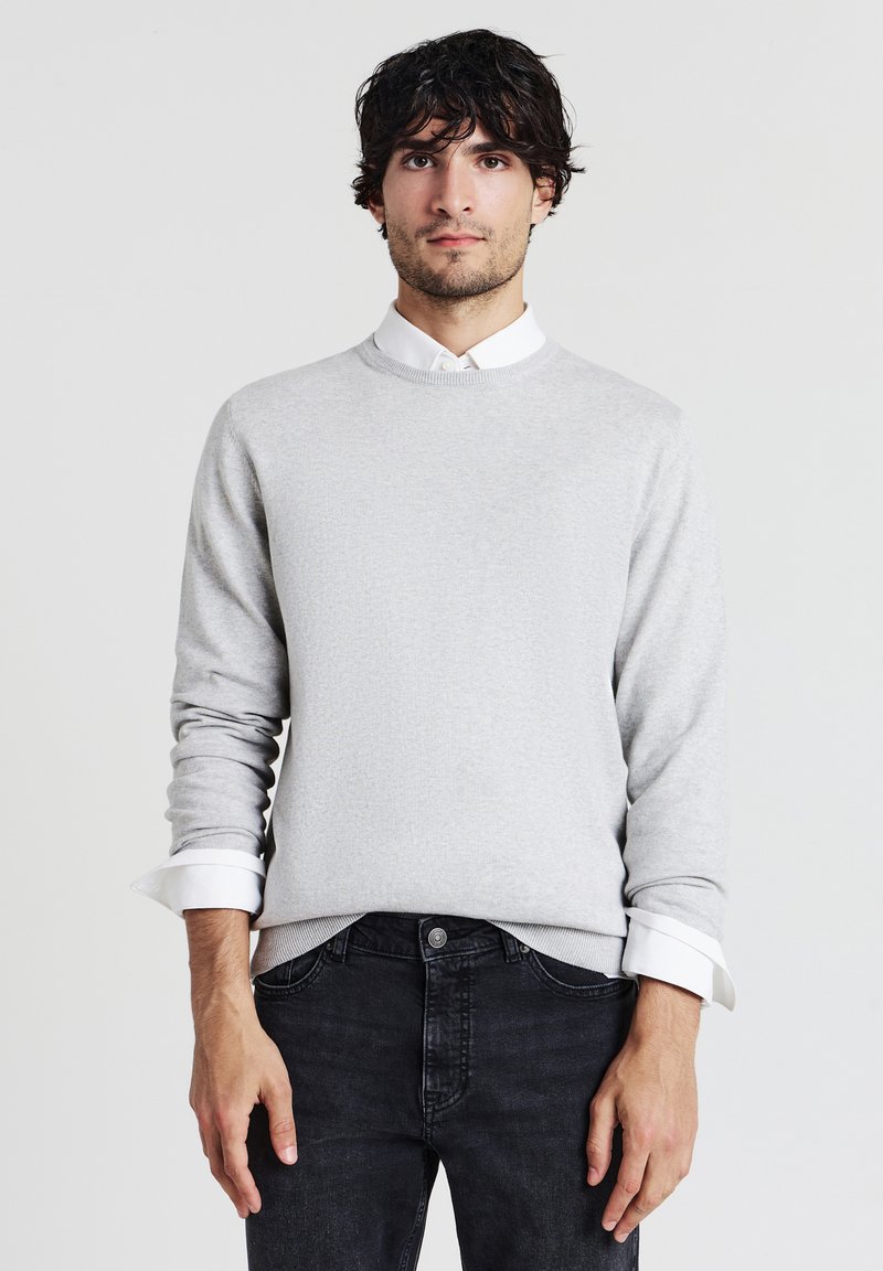 Junger Mann mit dunklem, lockigen Haar, der einen hellgrauen Pullover über einem weißen Hemd und schwarzen Jeans trägt, vor einem schlichten weißen Hintergrund stehend.