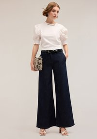 Bluza albă cu mâneci bufante, decolteu brodat, pantaloni evazați din denim închis, cu talie înaltă, curea neagră și poșetă cu imprimeu de șarpe.