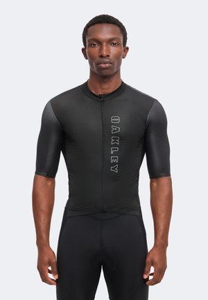 Mand iført sort cykeltrøje med korte ærmer med teksturerede ærmer og "Oakley" logo lodret på fronten, stående mod ensfarvet baggrund.